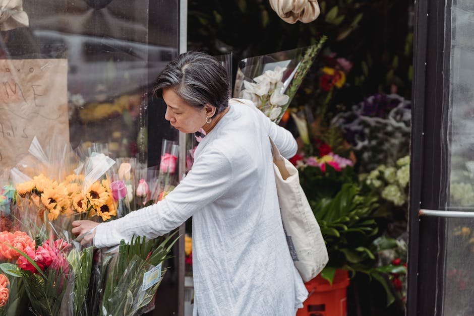 A woman selecting fresh floral bouquets at a flower shop entrance, with arrangements of pink and peach roses, yellow daisies, and purple lisianthus wrapped in clear plastic, displayed on black shelvin