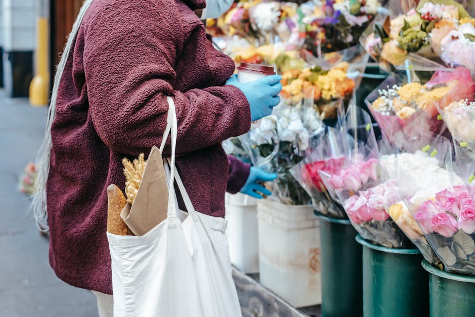 A person wearing a maroon, textured fleece jacket and blue gloves stands at a flower stall on East Street Market in Walworth. They are holding a small container of floral arranging supplies in one han